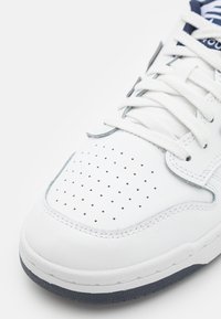 Baskets blanches en cuir avec un orteil perforé, des lacets blancs et une semelle en caoutchouc bleu foncé. Coutures d'accent visibles le long des bords.