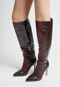 Botas hasta la rodilla en cuero marrón oscuro con estampado de cocodrilo, con puntera puntiaguda, tacón de aguja y detalle de cremallera lateral. Textura suave en toda la superficie.
