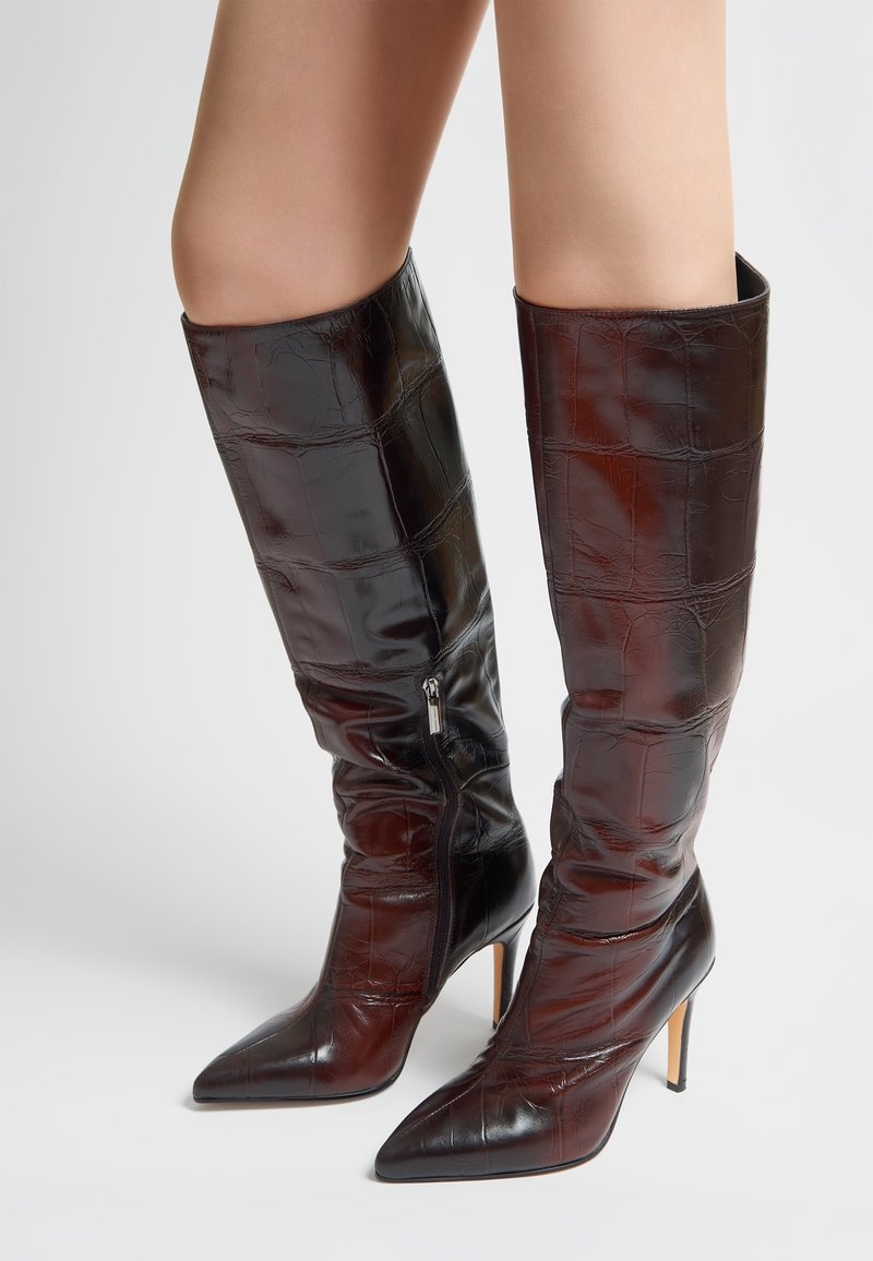 Botas hasta la rodilla en cuero marrón oscuro con estampado de cocodrilo, con puntera puntiaguda, tacón de aguja y detalle de cremallera lateral. Textura suave en toda la superficie.