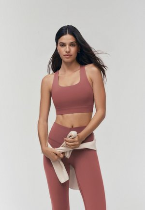Vrouw met lang donker haar, die een mauve sportbeha en legging draagt, knoopt een lichtbeige trui om haar middel tegen een effen achtergrond.