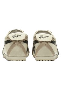 Par af beige og sorte sportssko med "Tiger" logo på hæltabberne, synlige syninger og teksturerede gummisåler set fra bagsiden.