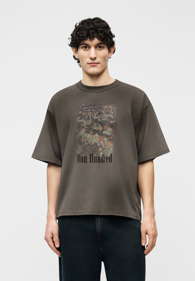 Νέος άνδρας με κοντά σγουρά μαλλιά φοράει υπερμεγέθη σκούρο γκρι μπλουζάκι με ξεθωριασμένο γραφικό πλήθους και κείμενο "Won Hundred," συνδυασμένο με σκούρο παντελόνι.