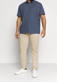 Marineblauw geruit overhemd met korte mouwen, beige chino's en witte sneakers. Het overhemd heeft knopen en een kraag; de chino's zijn opgerold bij de pijpen.