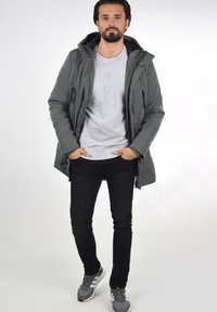Šedá zateplená bunda s kapucí, bočními zipovými kapsami, nošena přes šedé tričko, v kombinaci s černými slim-fit džíny a šedými teniskami.
