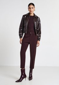 Femme portant une veste croppée brillante marron foncé, un pull côtelé, un pantalon tailleur et des bottines à talons aiguilles pointues, se tenant sur fond blanc.