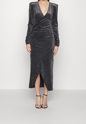 Mujer con un vestido cruzado de manga larga, oscuro y brillante, con escote en V profundo y abertura frontal, combinado con tacones negros de pulsera en el tobillo con adornos.