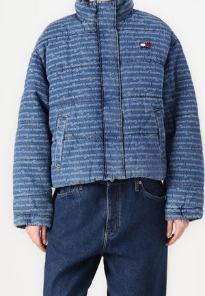 Veste matelassée bleue avec le texte répété "tommy jeans" et un petit logo Tommy Hilfiger, portée avec un jean denim bleu foncé.