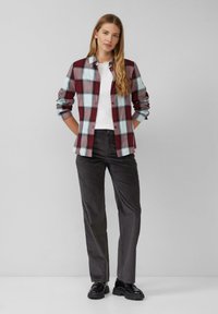 Chemise à carreaux rouge et bleu clair avec des manches longues sur une chemise blanche, associée à un pantalon gris foncé et des chaussures noires.