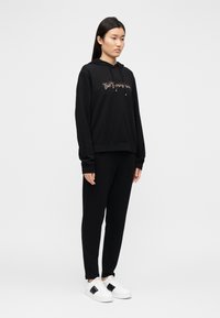 Μαύρο βαμβακερό φούτερ με κουκούλα και εμπρόσθιο γραφικό, συνδυασμένο με μαύρο παντελόνι και λευκά αθλητικά παπούτσια με μαύρες λεπτομέρειες.