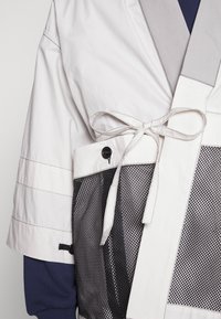 Lekki kimono w kolorze kremowym z szarymi akcentami, zapinany na materiałowy pasek, z siateczkową kieszenią. Teksturowana tkanina z kontrastującymi szwami.