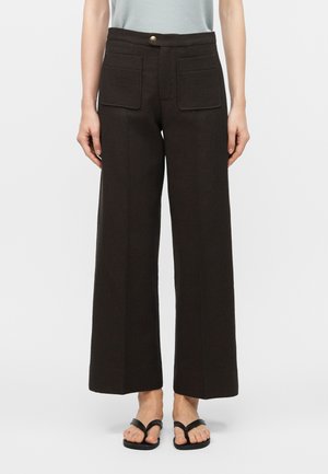 Személy magas derekú, bőszárú fekete nadrágot visel nagy első zsebekkel és fekete papucsot, sima háttér előtt állva.