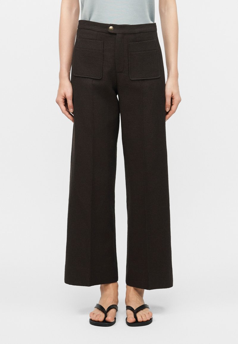 Άτομο που φοράει μαύρο παντελόνι ψηλόμεσο με φαρδιά μπατζάκια και μεγάλες μπροστινές τσέπες, καθώς και μαύρα σανδάλια τύπου σαγιονάρα, στέκεται μπροστά σε απλό φόντο.