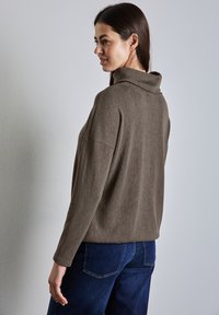 Brun, strukturerad tröja med hög krage, avslappnad passform och långa ärmar. Buren med blå denimbyxor, visad bakifrån.