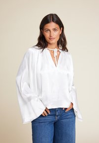 Hvit satinbluse med V-utringning og knytedetalj, med vide ermer og en avslappet passform, kombinert med blå denimbukser.