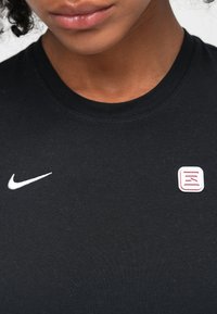 Crna pamučna majica kratkih rukava s okruglim izrezom, koja ima bijeli Nike logo i kvadratnu iglu s crvenim linijama na području prsnog dijela.