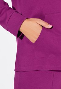 Fialová mikina s ribovanými manžetami, se přední kapsou. Hladký, měkký materiál s mírně volným střihem na spodním lemu.