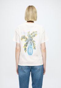 Tricou din bumbac crem cu un vas albastru cu design floral galben și verde pe spate. Croială lejeră și mâneci scurte.