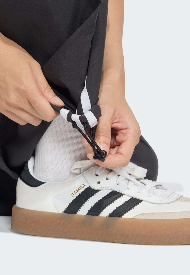 Hvide lærredssneakers med sorte striber, brun gummisål. Hænder justerer sort-hvid snor på bukser over en hvid sok.