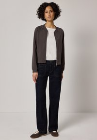 Frau mit lockigem Haar, trägt eine dunkle Jacke mit Reißverschluss, ein weißes T-Shirt, dunkle Jeans und braune Schnürschuhe, steht vor einem einfarbigen Hintergrund.