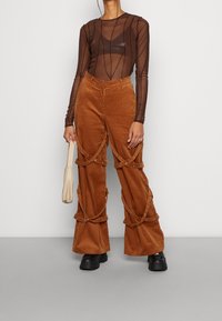Bruine corduroy broek met decoratieve banddetails, gecombineerd met een doorzichtige bruin lange mouwen top met constante verticale lijnen. Zwarte chunky schoenen.