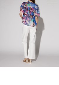 Blouse à fleurs bleue transparente avec des accents roses et marron, design superposé. Associée à un pantalon blanc évasé et des sandales à talons blocs dorés.