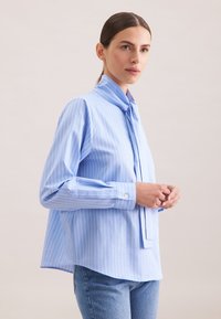 Hellbluse mit Streifen und Nackenbindchen, ausgestattet mit langen Ärmeln, Knopfmanschetten und einem abgerundeten Saum. Hergestellt aus glattem Stoff.