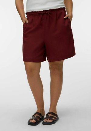 Person iført bordeauxfarvede snoreshorts med lommer og sorte sandaler med åben tå, stående mod en ensfarvet baggrund.