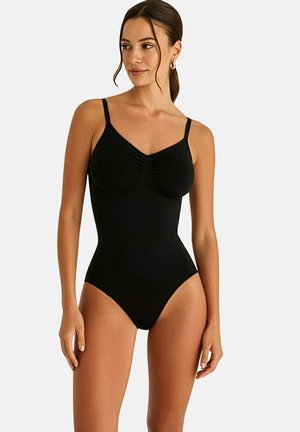 Femme portant un body noir une pièce avec des bretelles fines, debout devant un fond blanc uni.