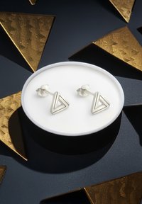 Silvertriangulära stiftörhängen med geometrisk design, visade på en cirkulär vit bas mot en mörk bakgrund med gyllene accenter.