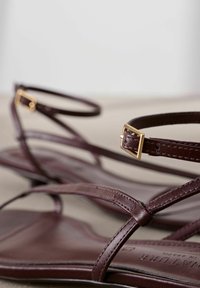 Sandalias de cuero marrón con correas finas, hebillas doradas y suela plana. Presentan una textura suave y un diseño minimalista.