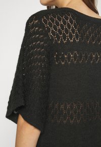 Femme portant un haut noir en maille à manches courtes avec des détails en ajouré à motifs dans le dos et sur les manches.