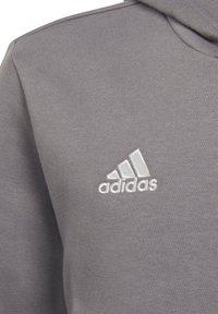 Grå hettegenser laget av myk stoff med ribbestrikkede mansjetter og et brodert hvitt Adidas-logo på venstre bryst.
