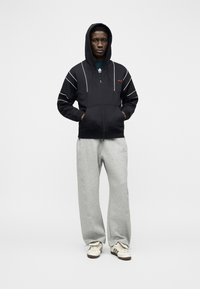 Άνδρας που φοράει μαύρη ζακέτα με φερμουάρ και λευκές γραμμές, καθώς και γκρι αθλητικό παντελόνι, στέκεται με τα χέρια στις τσέπες μπροστά σε έναν απλό φόντο.