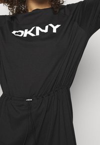 Svart klänning med en rynkad midja och dragsko. Har en stor vit logotyp fram. Mjuk, lätt tyg med korta ärmar.