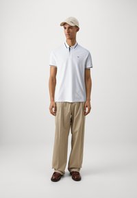 Camiseta polo blanca con ribete negro, pantalones anchos beige y una gorra ligera beige. Zapatos marrones completan el atuendo. Diseño minimalista y tela suave.