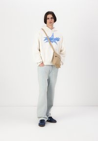Bolso de hombro beige, sudadera crema con gráfico azul, jeans de mezclilla azul claro y zapatillas navy, de pie contra un fondo blanco.