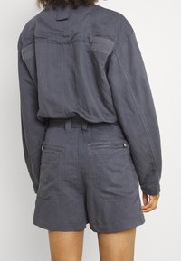 Camisa de lino gris con un corte relajado, que presenta detalles de velcro y dos bolsillo traseros en unos pantalones cortos a juego. Diseño casual y moderno.