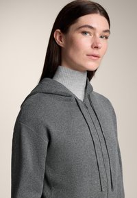 Hanorac gri cu glugă, cu design structurat, prevăzut cu fermoar frontal, șnururi și un gât înalt gri texturat dedesubt.