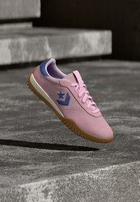 Ροζ αθλητικό παπούτσι με μπλε λογότυπο αστεριού και καφέ σόλα που αιωρείται πάνω από υφή σκυροδέματος, με θολά πέτρινα σκαλοπάτια στο φόντο.