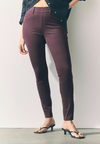 Dunne bordeauxrode broek gemaakt van rekbaar materiaal, met een hoge tailleband en zakken aan de voorkant, gecombineerd met zwarte sandalen met hakken en bandjes.
