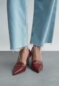 Røde lær høye hæler med spiss tå, med et gullakcent. Kombinert med lyseblå jeans med frynset kant.