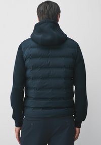 Veste matelassée bleue marine avec capuche, présentant une texture de tissu lisse, un design matelassé horizontal et des poignets élastiques, portée sur un pantalon sombre.