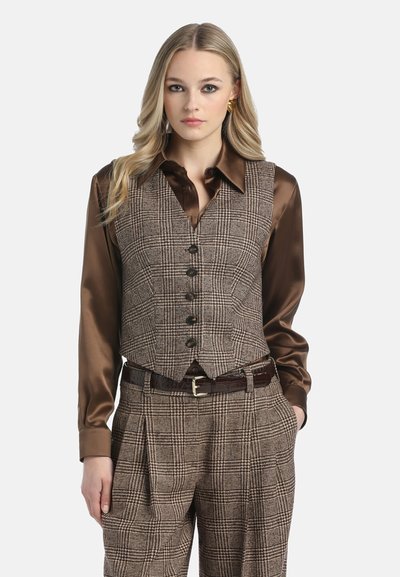 Smeđi karirani prsluk preko smeđe satenske košulje. Dizajn s V-izrezom s gumbima i kontrastnim teksturama. Model nosi smeđi remen i hlače s uzorkom.
