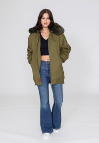 Olijfgroene bomberjack met een nepbont kraag, ritssluiting en twee zijzakken, gedragen over een zwarte top en wijde blauwe jeans.