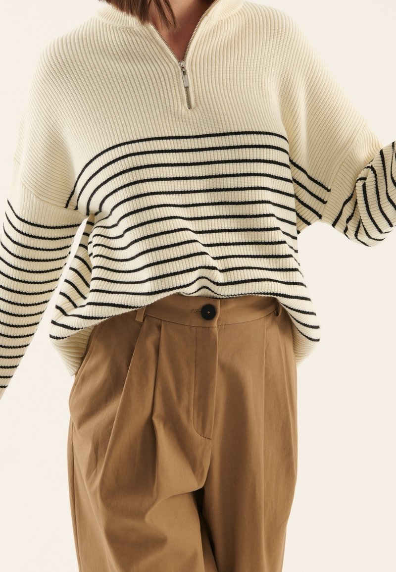 Femme porte un pull côtelé crème avec des rayures horizontales noires et un col montant zippé, associé à un pantalon plissé taille haute beige avec fermeture par bouton.