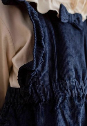 Tummansininen corduroy-haalarit, joissa on röyhelöreunat, yhdistettynä vaaleanbeigeen pitkähihanteeseen, jolla on pehmeä tekstuuri. Haalarissa on vyöltä koristeena oleva solmu.