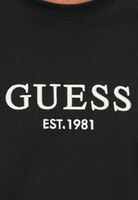 Čierna tkanina s bielym vyšitým textom "GUESS" nad menším textom "EST. 1981" v strede oblasti hrudníka.