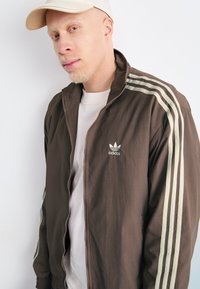 Brun glidelåsjakke med lysegrønne striper på ermene, lett materiale, adidas-logo på brystet, brukt over en lys t-skjorte.