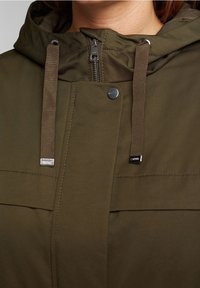 Chaqueta con capucha de color verde oliva, con cierre de cremallera en la parte delantera, cierre de botón a presión metálico y cordones planos. Cuenta con dos bolsillos en el pecho y una textura suave.