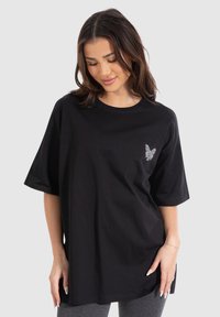Sort oversized T-shirt lavet af bomuld med korte ærmer, rund halsudskæring og et sølvfærge grafisk motiv på venstre bryst.